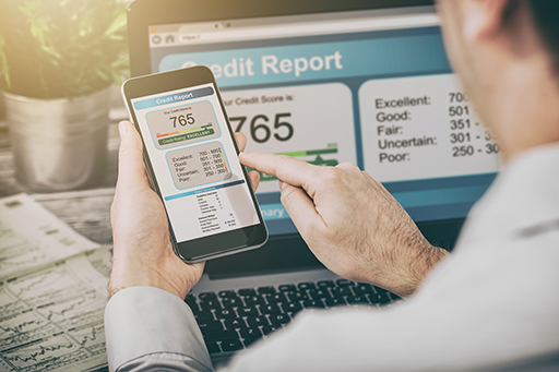The image shows a man using both a mobile ‘phone and a laptop to read their credit report. Their credit score is 765. Next to the laptop are some documents which have been extensively written on.