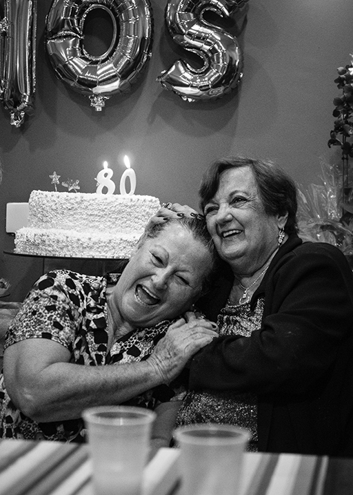 The figure is a photo of two elderly women with arms around each other at an 80th birthday celebration. In the background is a large cake with the number ‘80’ pinned on top of it.