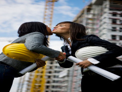 Photo of two women standing in their workplace and kissing on the cheek. They are both dressed in a smart-casual style.
