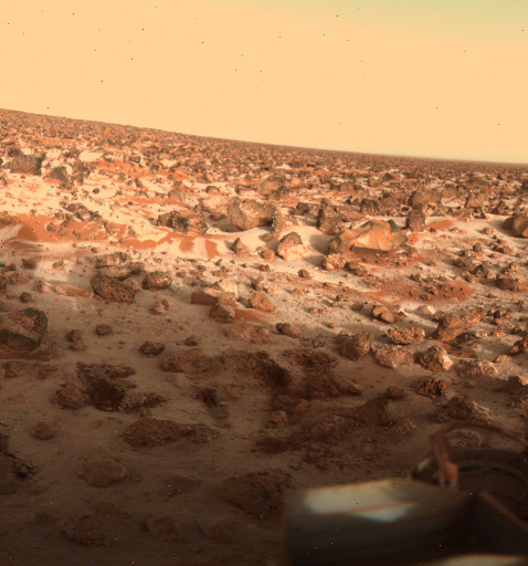 This is a photograph of the martian surface taken by the Viking 2 lander. The horizon is sloping from left to right. The sky is a pale orange colour. The surface is covered in red-brown dust with red-brown boulders of various sizes. The surface between the boulders has a dusting of white, which is water ice.