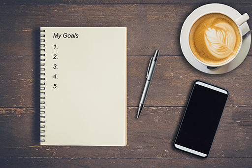 There is a notebook with the heading ‘My goals’ next to a pen, phone and cup of coffee.