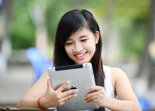 A photo of a young woman with long, dark hair, reading from a tablet computer. She is smiling.