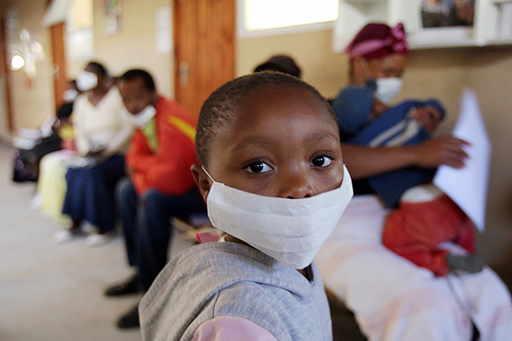 An image with a young boy wearing a medical mask, in the foreground; a patient in a South African tuberculosis clinic