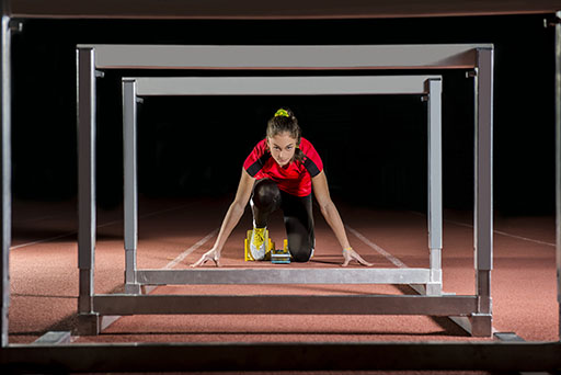 A female athlete wearing a red top is crouched ready to race in the starting blocks.
