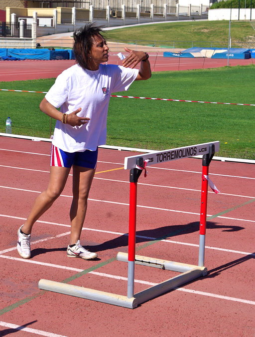 A woman in athletic gear stands on a racetrack in front of a hurdle.