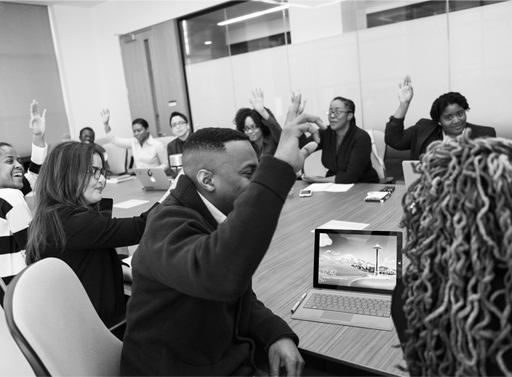 A photograph of people at a meeting. The people have their hands up in response to something.