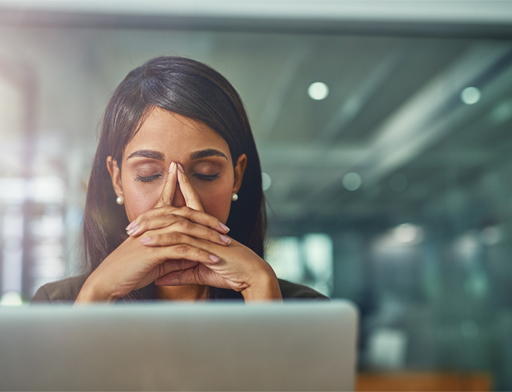 A photograph of a woman sitting at her desk covering her face with her hands.