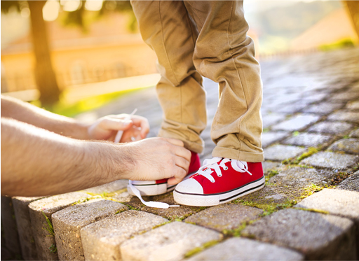 Image shows the hands of an adult man tying up the shoelaces of a young child