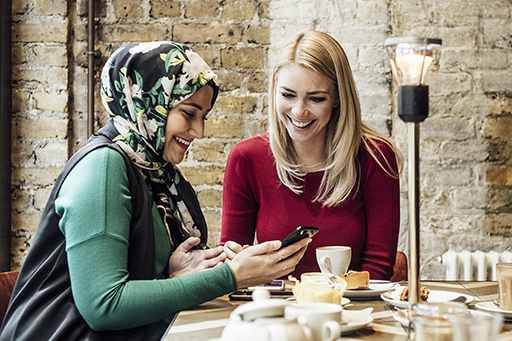 The image is a photo of two women in a café. One woman has her mobile phone in her hand and both women are looking at the screen.