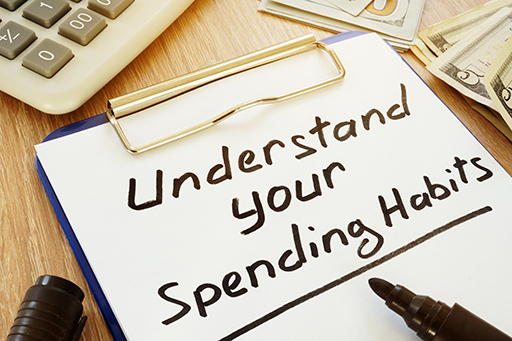 The figure shows a clipboard with a piece of paper inserted to it. On the paper is written ‘Understand your spending habits’. Around the clipboard are some bank notes and a calculator.