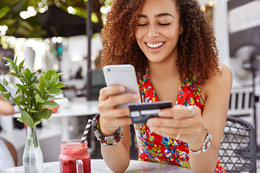 The figure is a photo of a young women sitting at a table in a café holding a credit card and a mobile ‘phone as she makes an online transaction.