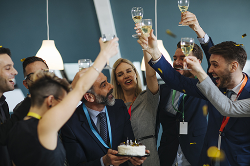 The figure is a photo of a retirement party at a workplace. The man retiring holds a cake. His colleagues are raising their glasses to him.