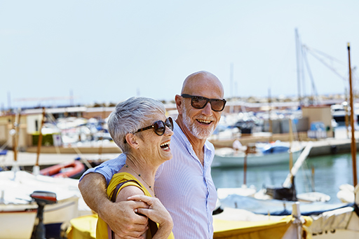 The figure is a photo of a (younger) retired male and female couple, smiling and enjoying themselves whilst walking around a harbour. Boats can be seen in the background.
