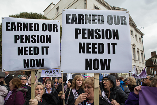 The figure is a photo of women demonstrating about the changes to the UK state pension that have disadvantaged women. Two large placards are displayed, each stating ‘earned our pension need it now’.