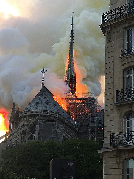This is a colour photo of Notre-Dame cathedral taken from the north-east. The fire on the roof of the church has completely overtaken the spire and some scaffolding arranged around it. The beams of the spire are visible through the smoke and flames.