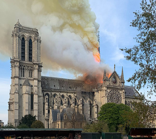 This is a colour photo taken of Notre-Dame Cathedral from the south. On the left is one of the large square belfries. To the right of centre, the roof above a stained-glass rose window is covered in bright orange flame. The flame has also spread part way up the spire behind the window and is producing plumes of smoke which fill the sky.
