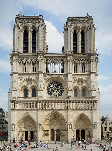 This is a colour photograph of Notre-Dame Cathedral from the public square to the west. Notable features of the cathedral include the two large square belfries on each corner, a large stained-glass rose window, and three large arches on the ground each containing the doors to the cathedral. In the foreground there are many people in the square who are probably tourists.