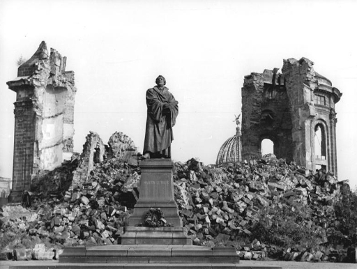 This is a black-and-white photograph taken shortly after the bombing of Dresden in the Second World War. It shows a statue of Martin Luther, made of black marble, standing on a stone plinth. Behind the statue there is a huge pile of rubble and two unsupported sections of wall belonging to the now-destroyed Frauenkirche.