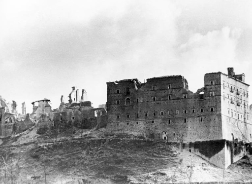 This is a black-and-white photograph taken in nineteen forty-four. It shows Monte Cassino Abbey after the bombing. On the right there is a square building which has several large craters in its roof and walls. To the left there is another connected building which is almost entirely ruined. All that remains are a few walls rising unsupported from the rubble.