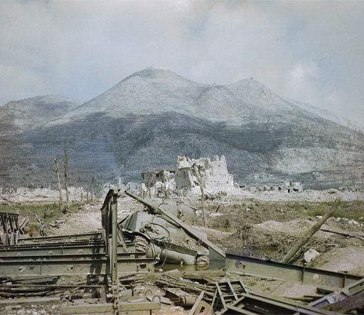 This is a colour photograph taken in nineteen forty-four. In the foreground there is a pile of metal wreckage and a broken-down green Sherman tank. In the mid-ground there is a churned-up field before a large area of bombed white buildings, standing entirely in ruins. In the distance there is a large forested hill, with Monte Cassino Abbey on top of the peak.