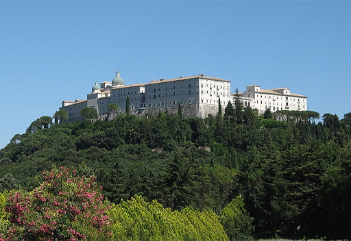 This is a colour photograph of Monte Cassino Abbey taken from a nearby hill in two thousand and seven. In the foreground there is a small forest, covering a steep incline. The Abbey rises from the trees in the centre of the photo. It is a large white building made of several cuboid buildings connected together with many small rectangular windows. Towards the left of the complex there are two small bluish domes on the roof.