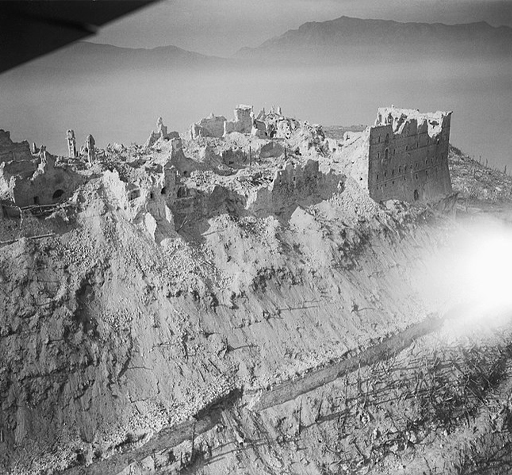 This is a black-and-white aerial photograph taken in nineteen forty-four. It shows the ruins of Monte Cassino Abbey from above. The building complex is set into the peak of a hill, which looks entirely bare due to the bombing. The buildings lie in almost complete ruins. No roofs remain; only scattered sections of walls stand above the rubble.
