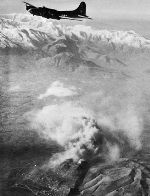 This is a black-and-white aerial photograph taken in nineteen forty-four. At the top there is a Boeing B-17 aeroplane (Flying Fortress) silhouetted against a snow-capped mountain range in the distance. The bottom of the photo shows an aerial perspective of the town of Cassino and the hill on which the abbey stands. The peak of the hill is obscured by a huge cloud of smoke – the result of a recent bombardment.