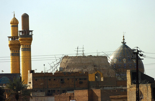 This is a colour photograph of the Al-Askari Mosque in Iraq, taken in two thousand and six. On the left it shows two golden minarets of the mosque rising above some buildings in the foreground. In the centre are the remains of the mosque’s dome, now missing its top half and covered in broken metal bars.