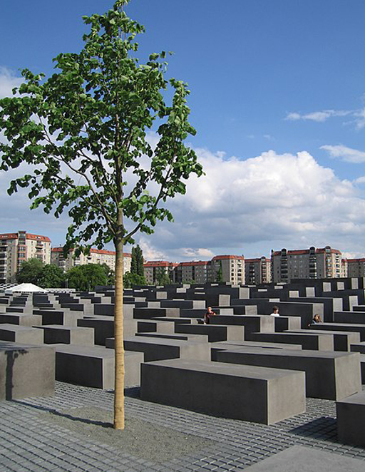 This is a colour photograph of the Holocaust memorial in Berlin. Countless grey concrete cuboids are arranged in rows, stretching off into the distance. The cubes are each about a metre wide and two or three metres long but varying in height. Some are close to the ground, while others are taller than head-height. In the distance there are some apartment buildings and in the foreground a single tree with green leaves.
