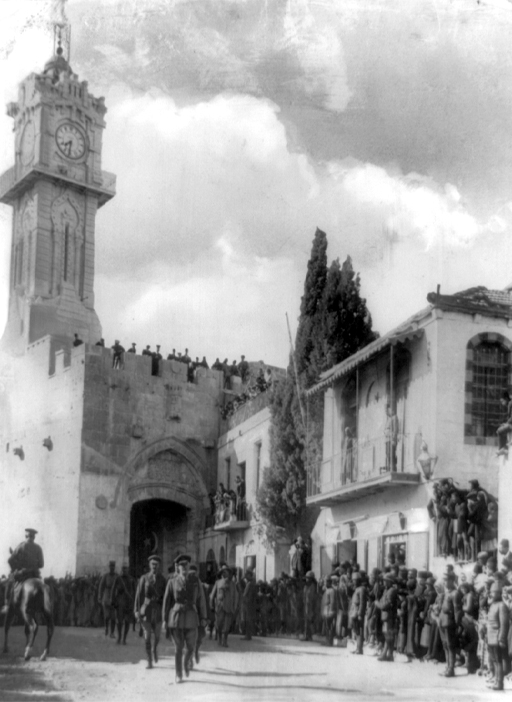 This is a black-and-white photograph taken in nineteen seventeen. It shows a large stone archway in Jerusalem, with an attached clock tower. In the foreground there is a street with a crowd of people and soldiers. Walking down the street towards the camera and from the archway is a group of soldiers with General Allenby at the front.