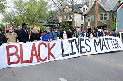 This is a photograph of a group of people holding up a banner which says ‘Black Lives Matter’.