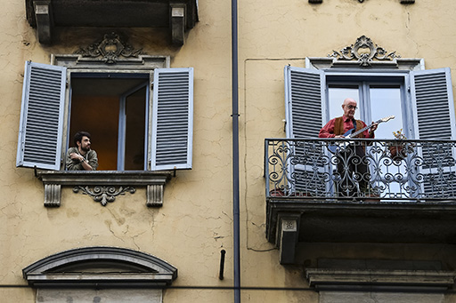 This is a photograph of a man standing on a balcony, playing the guitar. His neighbour looks on.