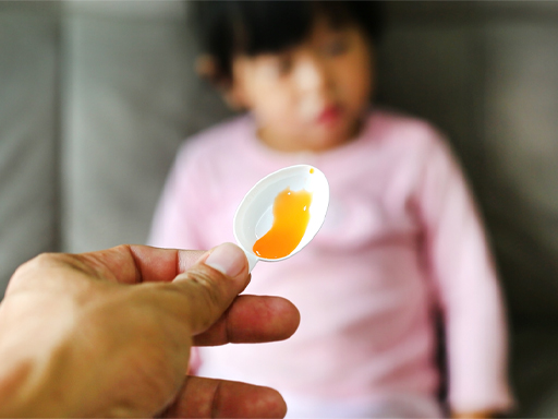 A photograph of an adult’s hand holding a spoonful of medicine, with a child in the background.