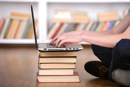 The image shows a person using a laptop which is sitting on top of a pile of books.