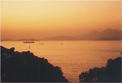 This photograph shows a sunset scene, looking over a body of water. The hue of the image is yellow and hazy.