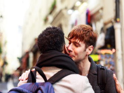 Photo of two young men kissing cheeks, each holding the other’s arm.. They are both dressed casually, one is wearing a cream-coloured coat, the other a black coat.