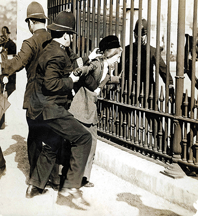 This is a black and white photograph of a woman in early twentieth-century dress holding on to railings, while a policeman tries to pull her away.