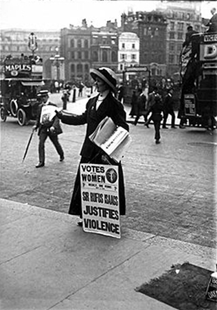 This is a black and white photograph of a woman in early twentieth-century dress standing at a kerb, facing on to the pavement. She has a placard resting against her leg and is holding a pile of leaflets under her left arm and holding one out in her right hand.