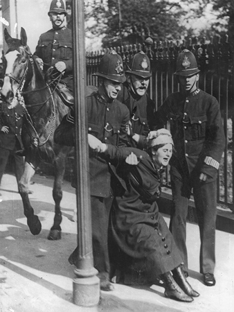 This is a black and white photograph of a woman in early twentieth-century dress being held from behind by three policemen. She appears to be struggling to break free. A fourth policeman sits on a horse behind the action, looking on.