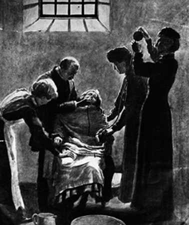 This picture in black and white shows a woman restrained in a chair, surrounded by four people – a man and three women. One of the women is pouring something into a tube which leads to the restrained woman’s nose. The other three people are holding the woman down.