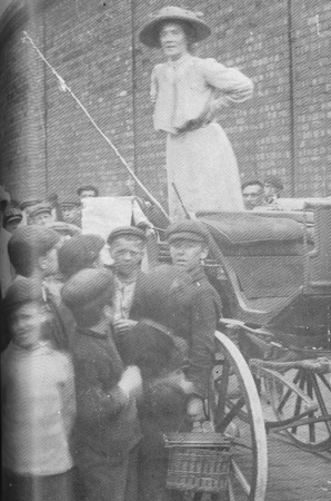 The black and white photograph shows Ada Nield Chew in hat and day-dress animatedly addressing a crowd during the Crewe by-election of 1912. She is standing high up on the back of a hansom cab and is surrounded by a throng of working-class men, whose cloth caps are very evident, and young working-class boys, again wearing cloth caps.