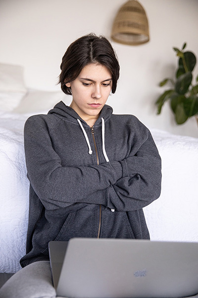 An image of a young person leaning against a bed with a laptop on their lap, arms folded and looking at the laptop.