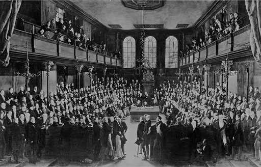 This black and white image depicts ranks of frock-coated men seated facing each other on either side of a large hall. Observers stand on balconies above them, which recede towards three high windows at the far end of the room.