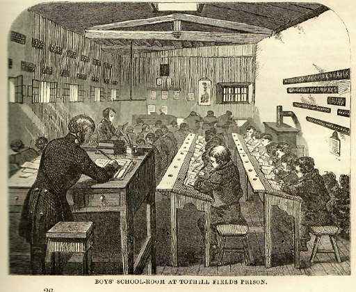 This is a black and white illustration of several rows of boys seated close together at desks in a long, narrow schoolroom. Their heads are bent as they write in their notebooks. They are supervised by two schoolmasters: one standing in the left foreground appears to be marking schoolwork; the other reads aloud from an open book.