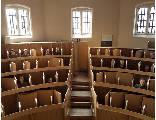 This colour photograph depicts a semi-circle of tiered pews, divided by a central aisle, beneath three windows in a rear wall. Seated boys occupy a few of the pews. They appear to be wearing a grey uniform, and are separated from each other by hinged wooden panels.