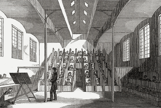 This black and white illustration depicts a high-ceilinged room, with about forty boys seated in tiered cubicles against the rear wall. In the left foreground stands a schoolmaster, pointing with a stick at a board on an easel. A few more boys are just visible at two rows of open desks along the side walls.