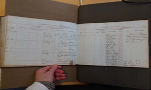 This is a colour photograph of an open record book held up against a screen. Handwritten entries have been made in a series of headed columns.