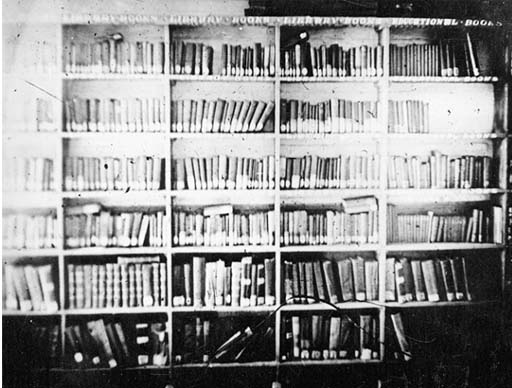This is a black and white photograph of a range of tall bookshelves, with a reading lamp in the foreground. The books are neatly arranged, with the larger ones on the lower shelves. All of them have white labels at the base of their spines.
