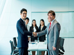 Two businessmen shake hands formally in front of a table in a meeting room, with other colleagues standing in the background.