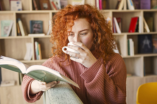 This is an image of a person sat with an open book in one hand and sipping from a mug in the other hand, reading.